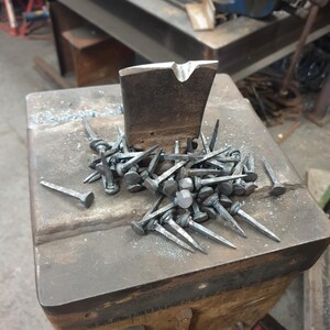 Pu&ograve; includere: Primo piano di un mucchio di chiodi forgiati a mano su un'incudine di metallo. I chiodi sono in acciaio e hanno una punta appuntita e una testa piatta. L'incudine &egrave; in ferro e ha una superficie ruvida.