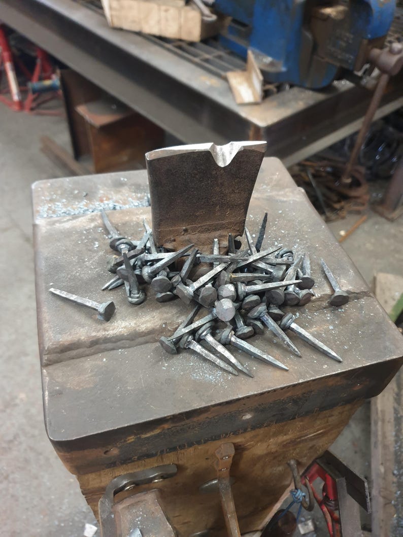 Pu&ograve; includere: Primo piano di un mucchio di chiodi forgiati a mano su un'incudine di metallo. I chiodi sono in acciaio e hanno una punta appuntita e una testa piatta. L'incudine &egrave; in ferro e ha una superficie ruvida.
