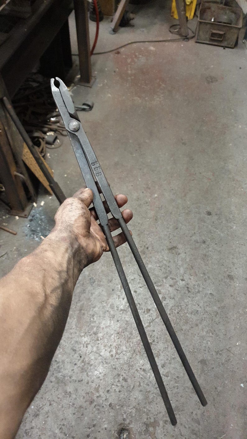 Może przedstawiać: Szczypce kowalskie z długimi uchwytami i chwytającą głowicą. Metalowe narzędzie trzymane jest w dłoni, ukazując otoczenie warsztatu. Szczypce mają wytłoczone litery "SLB". Narzędzie służy do trzymania gorącego metalu.