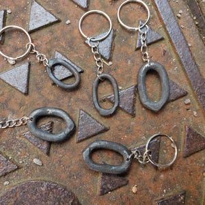 Pode incluir: Vários chaveiros com pingentes ovais de metal cinza escuro são exibidos. Cada chaveiro tem um anel prateado e uma corrente curta que se conecta ao pingente. Os chaveiros estão dispostos em uma superfície texturizada marrom enferrujado com formas triangulares.