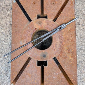 Może przedstawiać: Zardzewiałe metalowe narzędzie kowalskie z okrągłym środkiem i kątowymi wycięciami. Na narzędziu spoczywa para metalowych szczypiec. Narzędzie wydaje się być używane do kształtowania metalu. Og&oacute;lny kolor to brązowy z czarnymi akcentami.