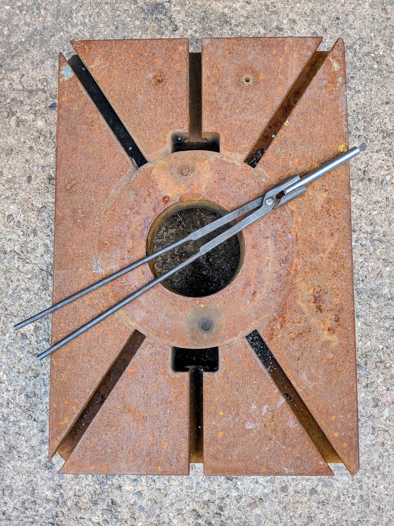 Może przedstawiać: Zardzewiałe metalowe narzędzie kowalskie z okrągłym środkiem i kątowymi wycięciami. Na narzędziu spoczywa para metalowych szczypiec. Narzędzie wydaje się być używane do kształtowania metalu. Og&oacute;lny kolor to brązowy z czarnymi akcentami.