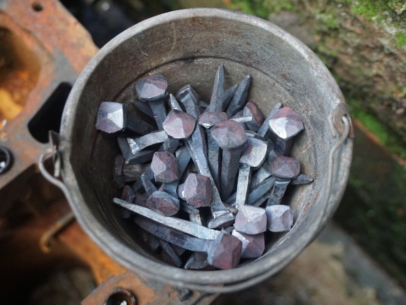 May include: A metal bucket filled with a collection of hand-forged nails with square heads. The nails are a dark grey colour and have a rustic appearance.