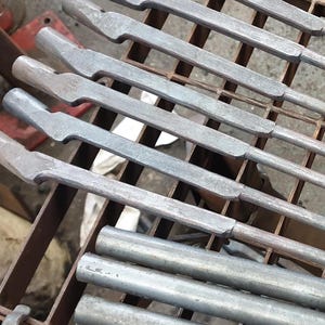 Może przedstawiać: Zbliżenie kilku metalowych narzędzi o teksturowanym, szarym wykończeniu, ułożonych na metalowym stojaku. Narzędzia mają charakterystyczny kształt z płaskim, kątowym końcem i cylindrycznym uchwytem. Obraz sugeruje warsztat lub środowisko przemysłowe.