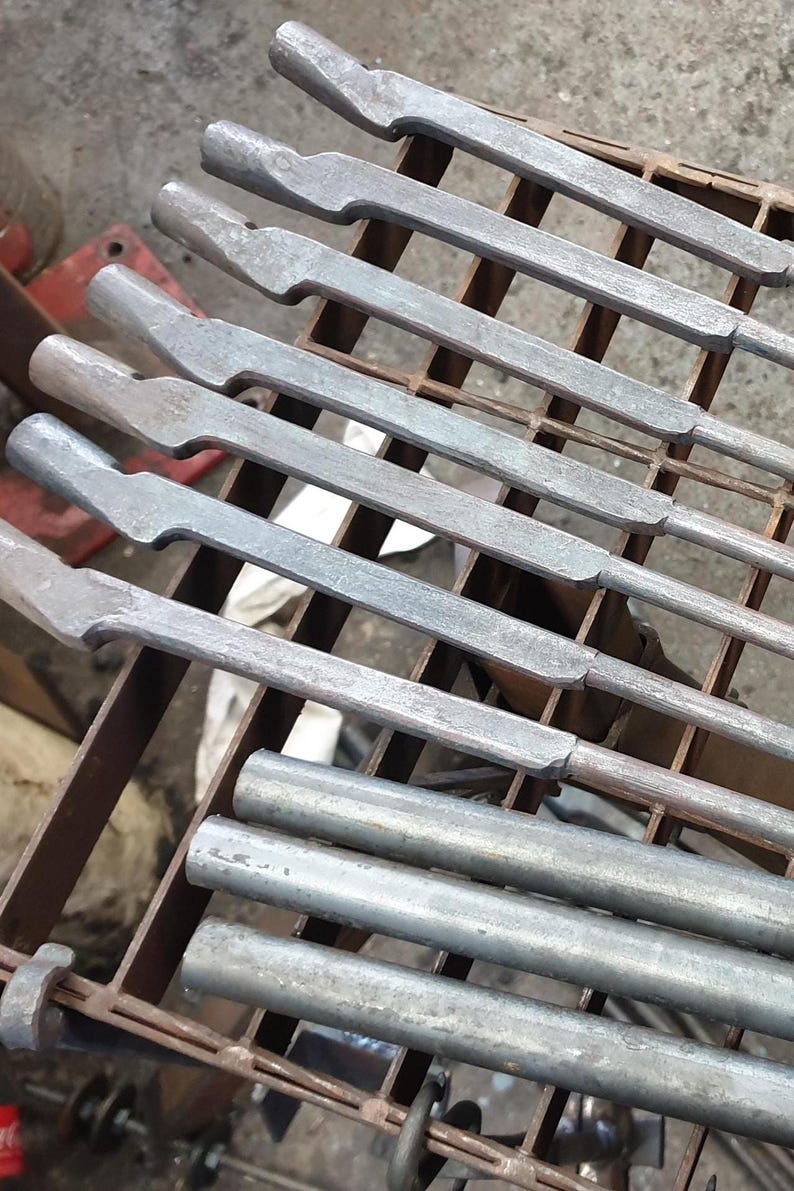 Może przedstawiać: Zbliżenie kilku metalowych narzędzi o teksturowanym, szarym wykończeniu, ułożonych na metalowym stojaku. Narzędzia mają charakterystyczny kształt z płaskim, kątowym końcem i cylindrycznym uchwytem. Obraz sugeruje warsztat lub środowisko przemysłowe.