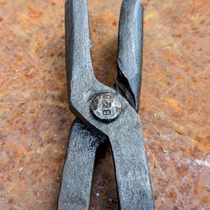 Może przedstawiać: Zbliżenie pary czarnych metalowych szczypiec, prawdopodobnie do kowalstwa lub obr&oacute;bki metali. Szczypce mają nitowane połączenie z wytłoczonymi literami "B&B". Tło to zardzewiała, teksturowana powierzchnia.
