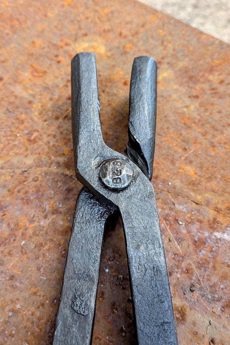 Może przedstawiać: Zbliżenie pary czarnych metalowych szczypiec, prawdopodobnie do kowalstwa lub obr&oacute;bki metali. Szczypce mają nitowane połączenie z wytłoczonymi literami "B&B". Tło to zardzewiała, teksturowana powierzchnia.