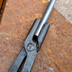 Może przedstawiać: Zbliżenie kleszczy kowalskich trzymających srebrny metalowy pręt. Kleszcze są czarne z nitami w punkcie obrotu. Tło to zardzewiała, teksturowana metalowa powierzchnia.