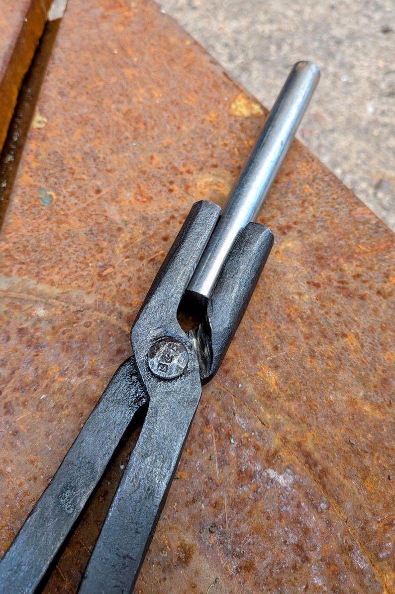 Może przedstawiać: Zbliżenie kleszczy kowalskich trzymających srebrny metalowy pręt. Kleszcze są czarne z nitami w punkcie obrotu. Tło to zardzewiała, teksturowana metalowa powierzchnia.