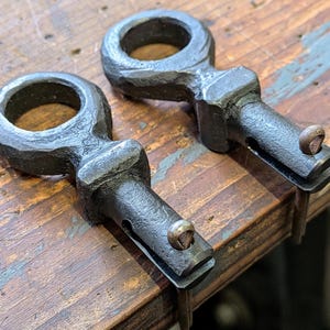 May include: Two dark metal, hand-forged brackets with a circular loop at the top and a slotted arm with a screw. The brackets are sitting on a weathered wooden surface. The metal has a textured, hammered appearance.