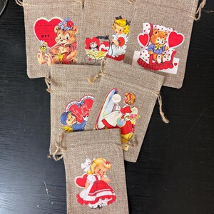 Könnte beinhalten: Sechs kleine Jutesäcke mit roten und weißen Valentinstags-Motiven. Die Bilder zeigen einen Teddybär, ein Mädchen in einem Kleid und eine Katze in einer Kutsche. Die Säcke haben Kordelzüge und sind perfekt für kleine Geschenke oder Mitbringsel.