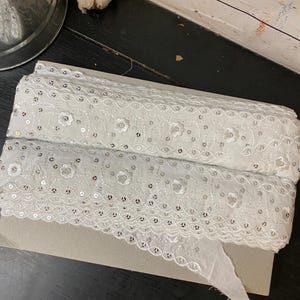 Può includere: Nastro di pizzo bianco con dettagli floreali e paillettes. Il nastro è arrotolato su una base di cartone. Il pizzo presenta un bordo smerlato, adatto a vari progetti di cucito e artigianato.