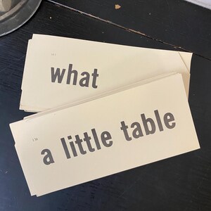 Könnte beinhalten: Zwei Vintage-Karteikarten mit schwarzem Text auf cremefarbenem Hintergrund. Eine Karte lautet "what" und die andere "a little table". Die Karten sind leicht angewinkelt, wobei die obere Karte die untere teilweise verdeckt. Der Hintergrund ist dunkel.