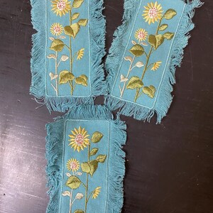 May include: Three rectangular teal coasters with embroidered sunflower designs. Each coaster features a yellow sunflower, pink buds, and green leaves on a stem. The edges are fringed.