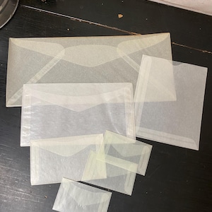 May include: A collection of translucent, off-white paper envelopes in various sizes. The envelopes are arranged on a dark surface, with a few overlapping. The paper has a subtle texture, and the envelopes have a classic, simple design.