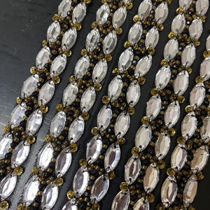 May include: Close-up of several rows of decorative trim. Each row features large, clear, oval-shaped rhinestones alternating with smaller, gold-colored beads. The trim is set against a black background, creating a striking contrast.