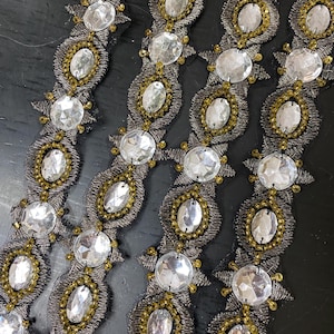 May include: Close-up of decorative trim featuring large, clear, oval-shaped rhinestones and smaller gold-coloured beads. The trim is set on a dark grey mesh backing with a metallic thread border, creating a sparkling, ornate design.