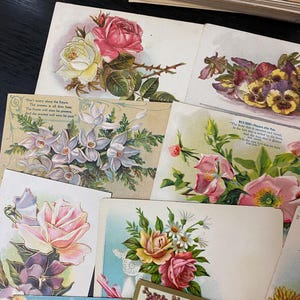 Puede incluir: Una colecci&oacute;n de postales florales vintage con rosas, pensamientos y otras flores en varios colores. Algunas tarjetas tienen texto, incluyendo citas y poemas. Las tarjetas est&aacute;n dispuestas sobre una superficie oscura.