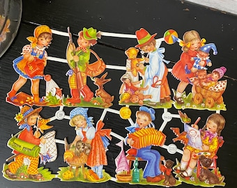 Puede incluir: Un conjunto de ocho muñecas de papel vintage que representan a niños realizando diversas actividades. Las muñecas están vestidas con ropa colorida y tienen rasgos detallados. El conjunto incluye una niña regando flores, un niño cazando, una niña jugando con un oso de peluche, una niña jugando con una muñeca, un niño tocando el acordeón, una niña pintando, una niña jugando con un perro y un niño jugando con un barco.