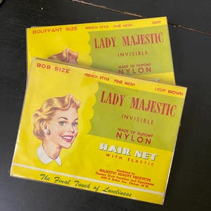 Puede incluir: Dos cajas de redecillas para el cabello vintage "Lady Majestic". Una es gris, la otra marrón claro. Ambas presentan una ilustración retro de una mujer con cabello rubio. El texto incluye "Invisible", "Made of Dupont Nylon" y "Hair Net with Elastic".