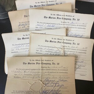 May include: A collection of aged paper documents secured with string. The documents are from The Marion Fire Company No. 10, featuring handwritten details like names and dates. The paper has a yellowed, vintage look.