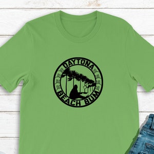 Peut inclure: Un t-shirt vert avec un graphique circulaire noir qui dit "Daytona Beach Bum" avec un palmier et une personne assise sur une chaise de plage.