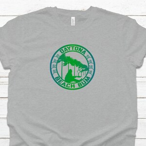 Peut inclure: T-shirt gris clair avec un motif circulaire vert et bleu indiquant "Daytona Beach Bum". Le motif représente des palmiers et une personne sur la plage. Le t-shirt est fait d'une matière douce.