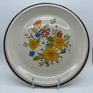 Op de afbeelding: Een rond keramieken bord met een gespikkelde gebroken witte basis en een donkerbruine rand. Het midden is voorzien van een kleurrijke bloemenillustratie met grote gele bloemen, kleinere witte madeliefjes, rode bloesems en blauwe blaadjes.
