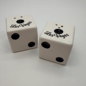 Könnte beinhalten: Ein Paar Salz- und Pfefferstreuer aus weißer Keramik in Würfelform. Jeder Würfel hat schwarze runde Punkte und den Schriftzug Las Vegas in schwarzer Schreibschrift auf der Oberseite. Die Streuer haben kleine Löcher an der Oberseite.