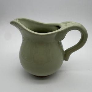 May include: A light sage green ceramic pitcher with a smooth, glossy finish. The vessel features a rounded body, a curved handle on the right side, and a prominent pouring spout. The base of the pitcher shows a thin band of unglazed terracotta clay.