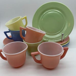 Könnte beinhalten: Eine Sammlung von Teetassen und Untertassen aus Milchglas im Vintage-Stil in Pastellfarben, darunter Hellgrün, sanftes Gelb, Hellblau, Pfirsich und Rosa. Die Tassen haben kantige Henkel und glatte, abgerundete Schalen, während die Untertassen rund sind.