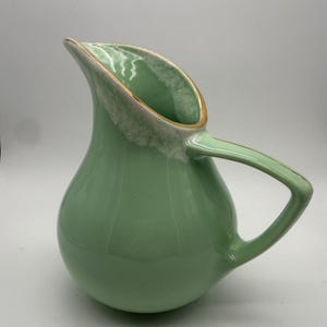 May include: A light mint green ceramic pitcher with a rounded body and a single angular handle. The rim features a distinctive white drip glaze effect that transitions into a smooth, glossy finish across the exterior of the vessel.