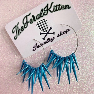 May include: A pair of silver hoop earrings with turquoise blue spikes. The earrings are displayed on a white card with the text "The Feral Kitten Jewelry Shop".