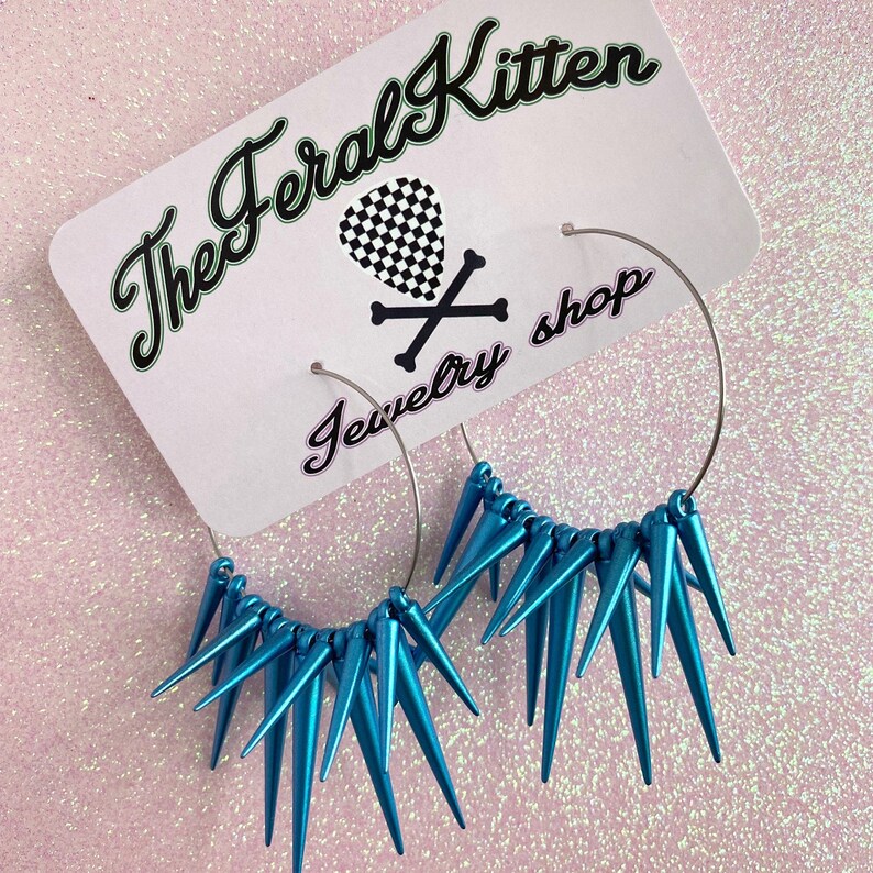 May include: A pair of silver hoop earrings with turquoise blue spikes. The earrings are displayed on a white card with the text "The Feral Kitten Jewelry Shop".