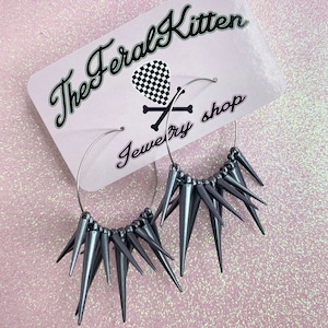 May include: A pair of silver hoop earrings with a row of silver spikes around the hoop. The earrings are on a pink background with the text "The Feral Kitten Jewelry Shop".