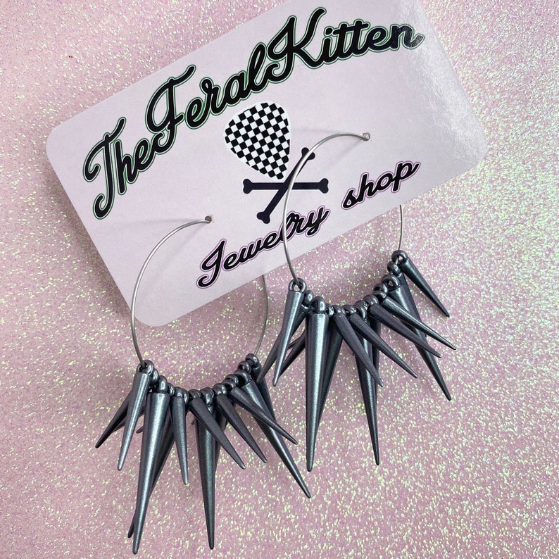 May include: A pair of silver hoop earrings with a row of silver spikes around the hoop. The earrings are on a pink background with the text "The Feral Kitten Jewelry Shop".