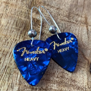 May include: A pair of blue Fender guitar pick earrings with silver hooks and beads. The picks have gold lettering that reads "Fender Heavy".