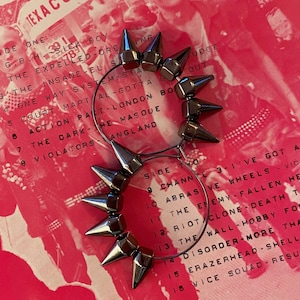 May include: A pair of silver hoop earrings with spikes. The earrings are on a pink background with text.