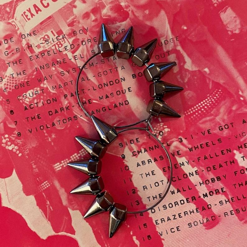 May include: A pair of silver hoop earrings with spikes. The earrings are on a pink background with text.