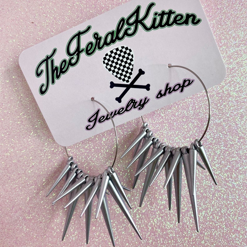 May include: Silver hoop earrings with silver spikes. The earrings are hanging from a white card with the text "The Feral Kitten Jewelry Shop".