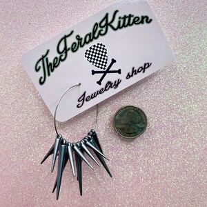 May include: Silver hoop earrings with a row of silver spikes hanging from the bottom. The earrings are displayed on a pink background with a business card that reads "The Feral Kitten Jewelry Shop".