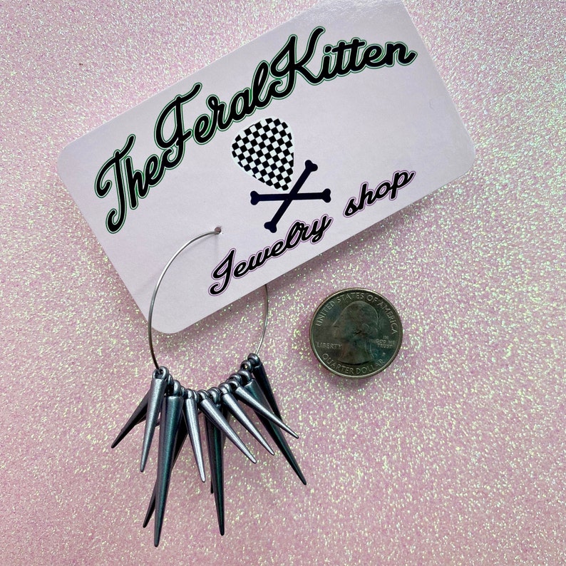 May include: Silver hoop earrings with a row of silver spikes hanging from the bottom. The earrings are displayed on a pink background with a business card that reads "The Feral Kitten Jewelry Shop".