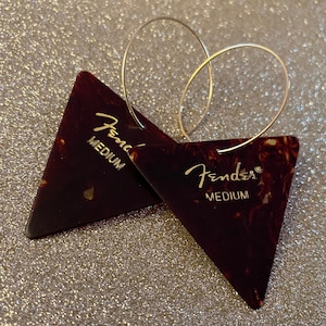 May include: A pair of silver hoop earrings with a brown and white Fender guitar pick attached to each hoop. The picks are labeled "Fender Medium".