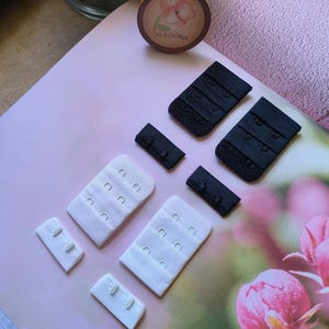 Puede incluir: Un juego de extensores de tirantes de sujetador en blanco y negro. Los extensores tienen múltiples cierres de gancho y ojo. Los extensores están sobre una superficie blanca.