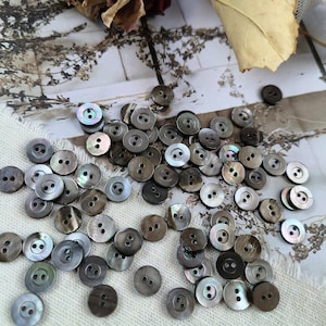 May include: A collection of approximately 50 dark grey, iridescent, two-hole buttons. The buttons are scattered on a white fabric surface.