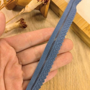 May include: A close-up of a blue elastic lace trim with a scalloped edge. The trim is held in a hand and is on a wooden surface.