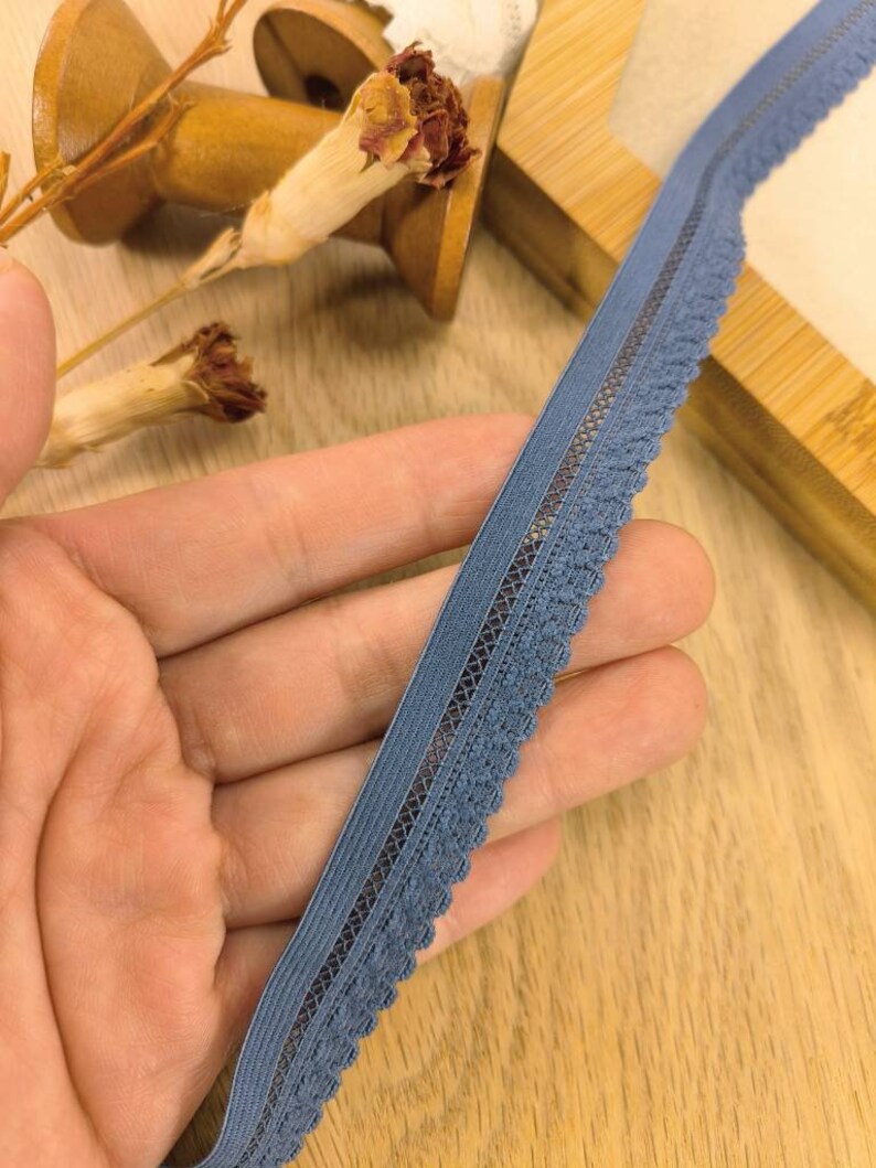 May include: A close-up of a blue elastic lace trim with a scalloped edge. The trim is held in a hand and is on a wooden surface.
