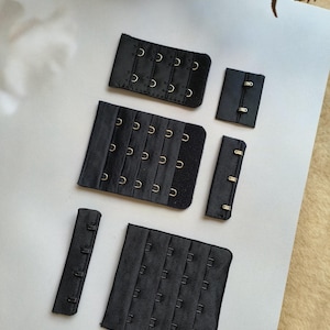 Puede incluir: Una colección de extensores de sujetador y cierres de corchete y ojal negros. Los extensores varían en tamaño y presentan múltiples juegos de corchetes y ojales. Los cierres son de tela y metal.