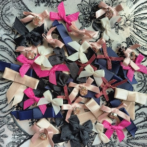 May include: A collection of small, colorful ribbon bows in various shades of pink, white, navy, and black. Some bows have a small, sparkly embellishment in the center. The bows are arranged on a white and black floral lace background.