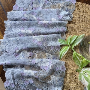 May include: Close-up of a folded, floral lace fabric in shades of gray, lavender, and white. The lace features a repeating pattern of roses. The fabric is displayed on a brown surface, with a plant in the background.
